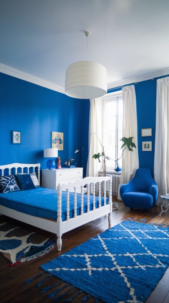 A cozy bedroom featuring blue walls, a traditional white bed frame, and a large white drum pendant light. A blue armchair and a blue and white patterned rug complement the room's main color scheme, contributing to its overall cozy and inviting ambiance.