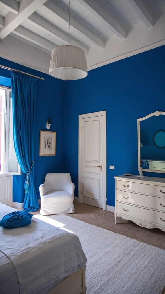 A cozy bedroom with vibrant blue walls and a white exposed beam ceiling. The room is appointed with a white bed, a white dresser, and a white armchair. A long blue curtain adds a touch of color, ensuring the space feels both lively and cozy.
