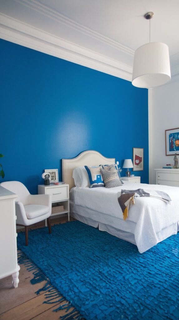 A cozy bedroom with a deep blue accent wall, contrasting with the remaining white walls and ceiling. The room is furnished with a white bed, white nightstands, and a large, textured blue rug, creating a relaxing and inviting cozy atmosphere.