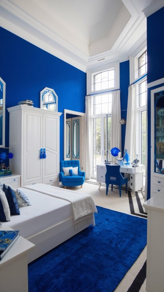 A spacious and cozy bedroom with bold blue walls and tall, white-framed windows letting in natural light. The room contains a white bed, a blue upholstered armchair, and a white desk with a matching blue chair, all situated on a deep blue area rug.