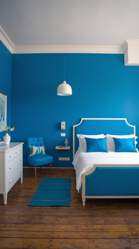 A minimalist and cozy bedroom with blue walls and a white bed featuring a blue upholstered headboard and base. The room includes white furniture, a blue accent chair, and a small, textured blue rug on the wooden floor, creating a simple yet inviting and cozy space.