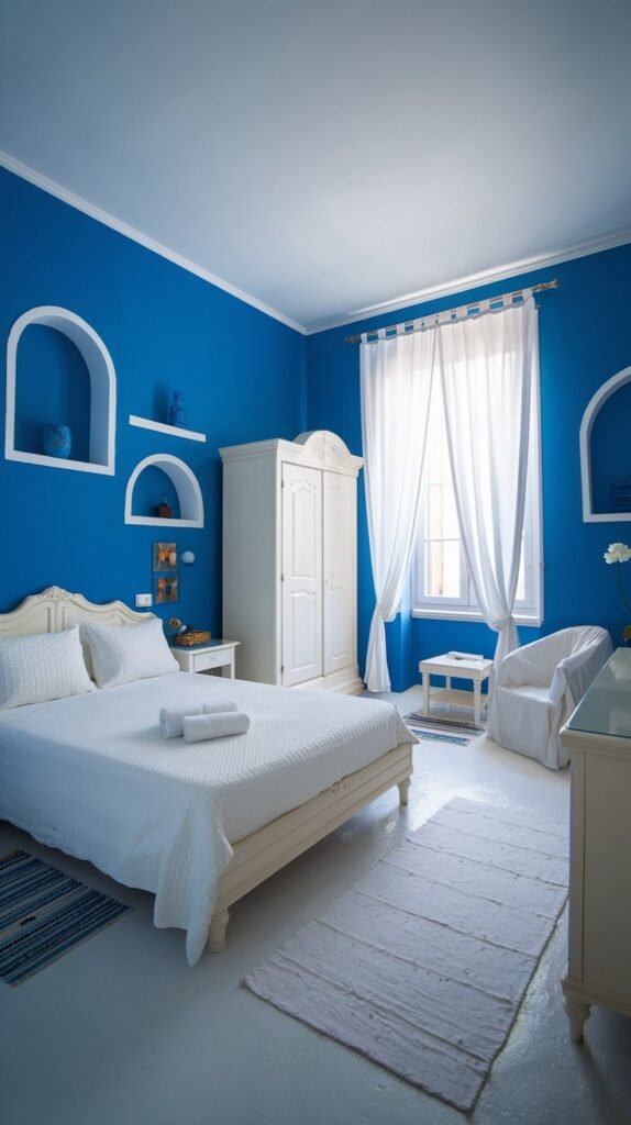 A serene and cozy bedroom with blue walls that feature arched wall niches for decor. The room is furnished with a white bed, a white wardrobe, and a white chair, complemented by white curtains on the window. A white rug with subtle blue accents on the side completes the peaceful and cozy look.