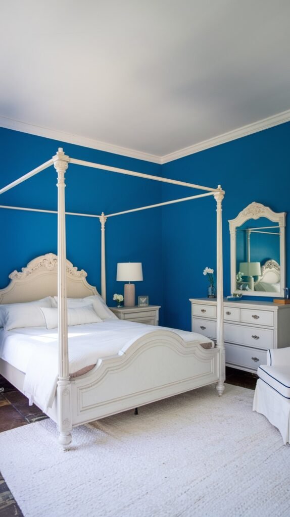 This cozy bedroom is painted a bright blue and features a stately white four-poster bed. The room is minimally furnished with white traditional furniture, including a nightstand and a dresser, along with a light-colored rug, creating a tranquil and cozy sanctuary.