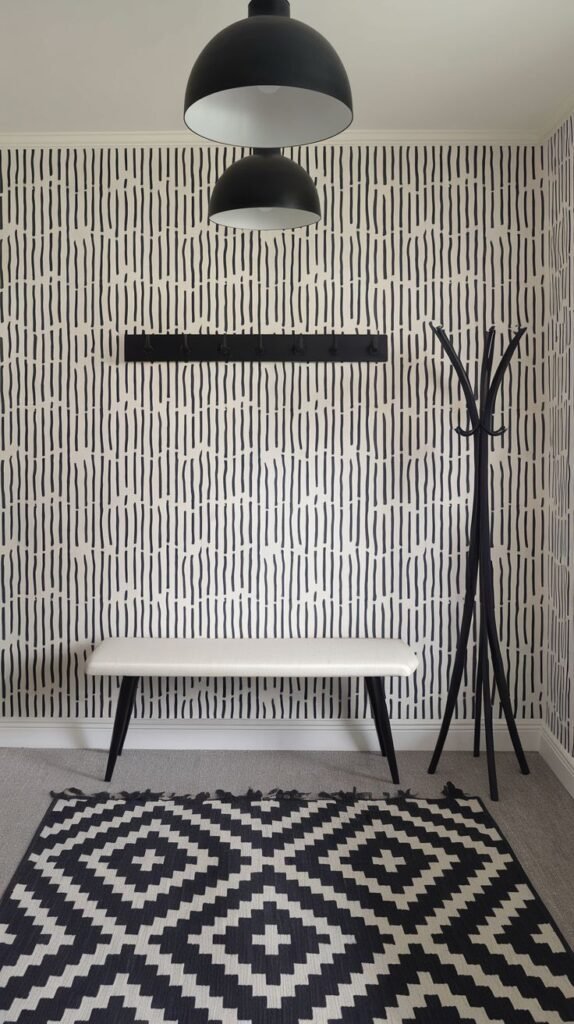 A dynamic Black and White entryway features a vertically striped wallpaper that creates movement, paired with a geometric patterned rug. Two large black dome pendant lights hang from the ceiling, and a sleek white bench with a black coat rack provides functional seating and storage.
