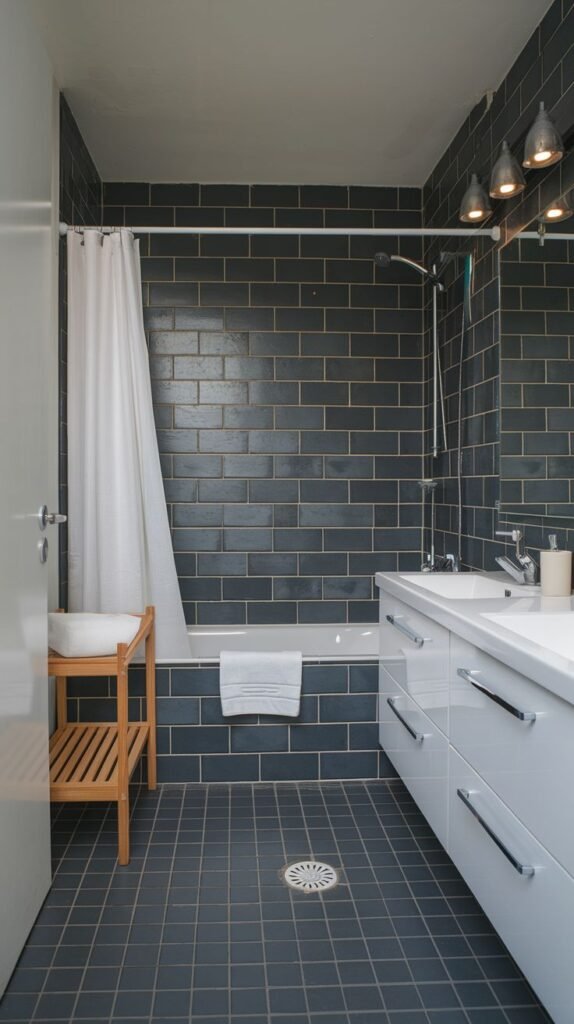 A compact bathroom with dark grey brick-patterned tiles on the walls and floor, a white bathtub with a shower curtain, and a white vanity with multiple drawers.