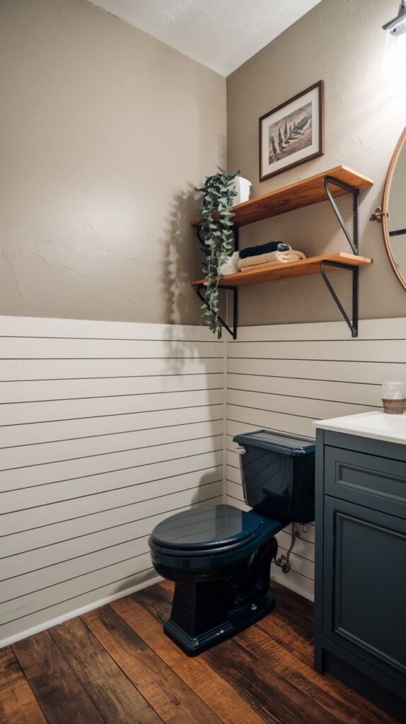 A modern rustic half bathroom with shiplap wainscoting on the lower walls and light beige painted upper walls. It features a dark blue toilet and a dark blue vanity with a white sink. Wooden floating shelves with a plant and decor are mounted above the toilet. The floor is dark wood planking.