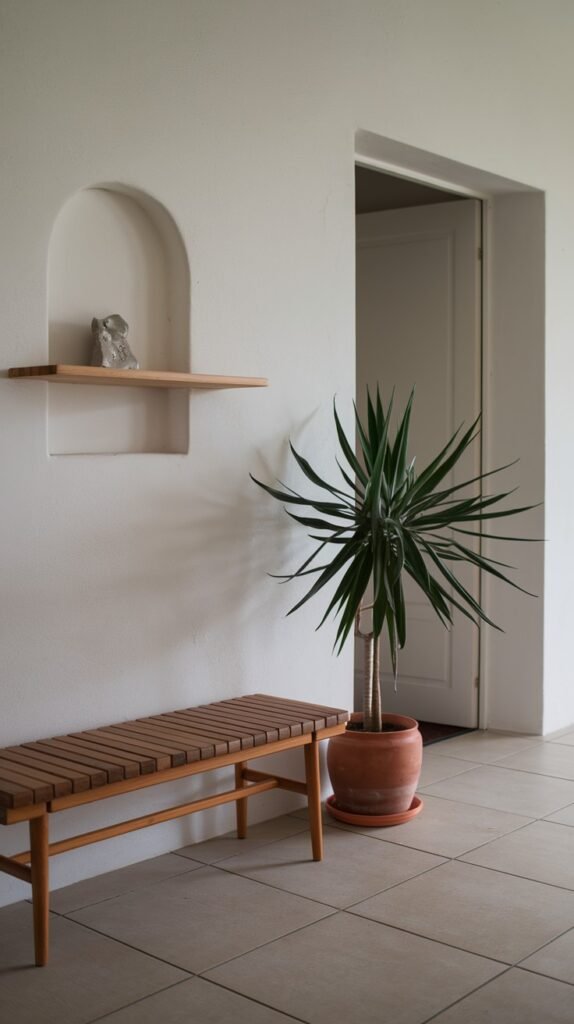 A simple entryway or hallway with a wooden slat bench and a large potted plant. The wall features a built-in arched niche with a floating shelf and a glass decoration, indicative of minimalist home decor.