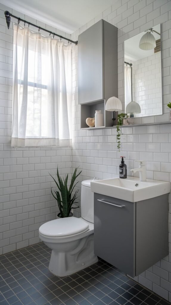 A small, modern bathroom showcasing white subway tiles, a grey floating vanity, and a matching overhead cabinet. It includes a toilet and a window with soft, sheer curtains. A plant sits on the floor and a smaller one on a shelf.