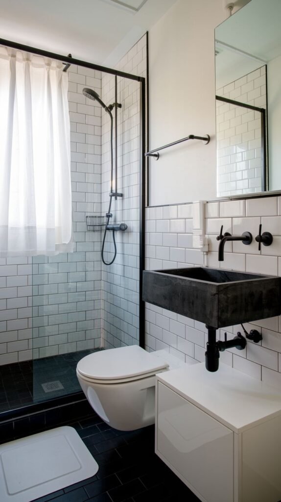 A small, modern bathroom with white subway tiles, a black tiled shower floor, and a black-framed glass shower screen. It includes a unique black concrete vessel sink mounted on a white cabinet and a wall-mounted toilet.
