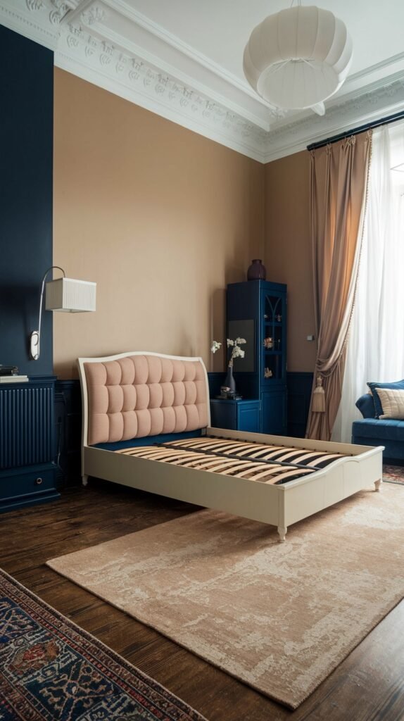 A bedroom with a mix of blue and beige tones. The room has a dark blue accent wall and beige painted walls. A bed with a light pink tufted headboard and a light-colored frame is featured. Dark blue furniture pieces, including a cabinet and a sofa, are also present. A light beige patterned rug covers the floor.