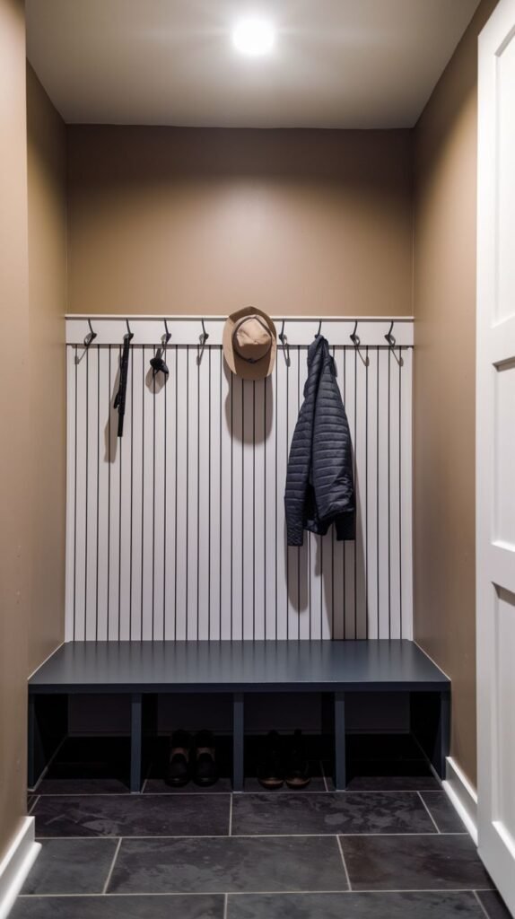 A small entryway mudroom with a modern design, featuring a dark gray bench with shoe storage cubbies. The wall behind the bench is covered in white vertical slat paneling with black hooks for hanging coats and a hat. The walls are light brown, and the floor is dark slate tile.