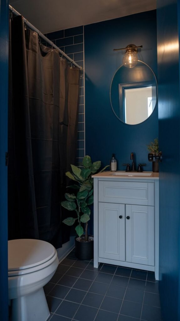 A tiny, moody bathroom with deep blue walls, a dark tiled floor, a white toilet, and a light gray vanity with a white sink. A black shower curtain and a round mirror complete the space.
