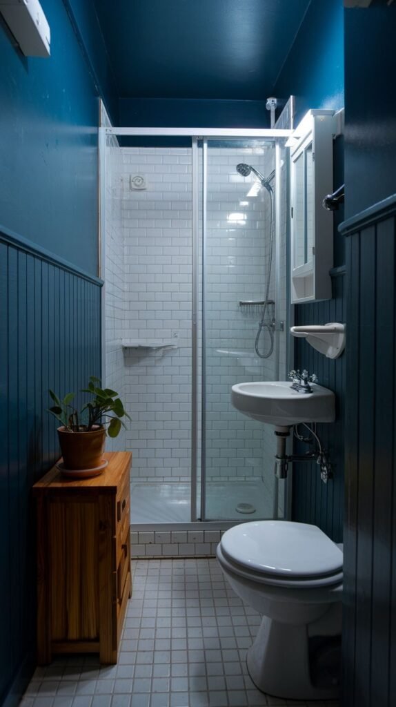 A tiny, moody bathroom with deep blue upper walls and natural wood paneling on the lower half. It features a white tiled floor, a white toilet, a small white wall-mounted sink, a glass-enclosed shower stall, and a wooden cabinet.