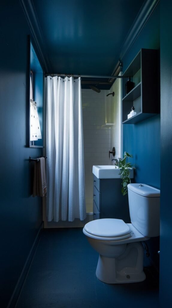 A tiny, moody bathroom with dark blue walls and a matching dark blue floor. It includes a white toilet, a small white wall-mounted sink, a shower with a white and gray striped curtain, a window, and a black open shelf.