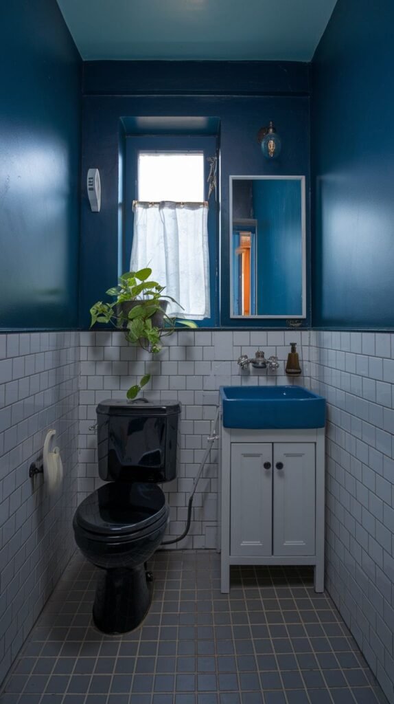 A tiny, moody bathroom with deep blue upper walls and white subway tiles on the lower half. It features a black toilet, a white vanity with a blue sink, a window with a sheer white curtain, and a dark tiled floor.