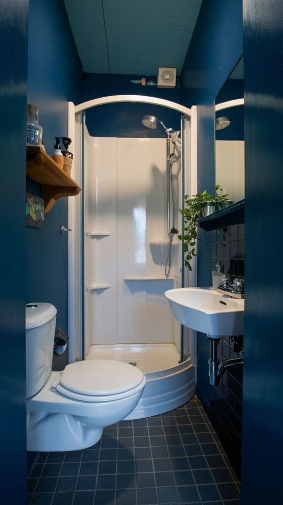 A tiny, moody bathroom with deep blue walls and a dark tiled floor. It features a white toilet, a small white sink, and a white curved shower stall.