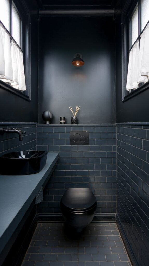 A tiny, moody bathroom with black walls, black subway tiles on the lower half, and a black tiled floor. It features a black wall-mounted toilet, a black sink basin on a gray countertop, and two windows with white curtains.