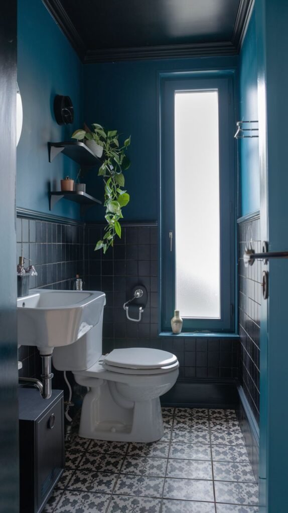 A tiny, moody bathroom with deep blue upper walls and black subway tiles on the lower half. It features a white toilet, a white sink, a tall frosted window, and a black and white patterned tiled floor.