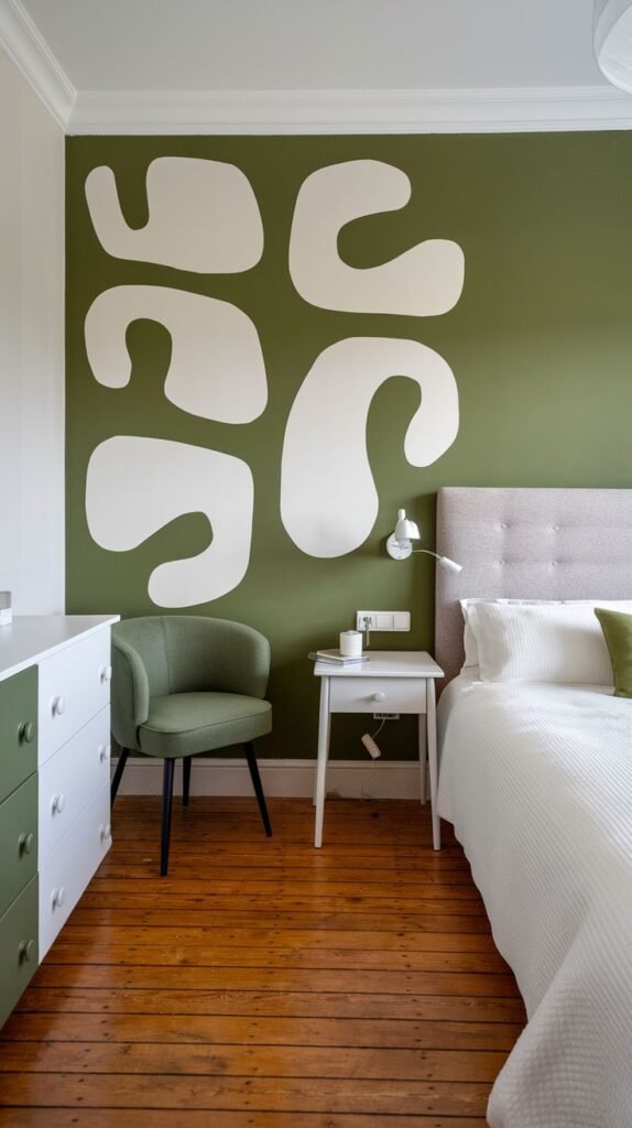 A warm green accent wall in a bedroom is adorned with large, free-form white organic shapes, creating a bold and artistic statement next to a simple bed and a green chair.