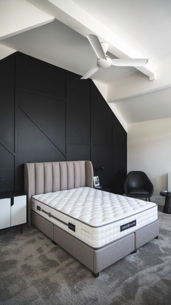 A bedroom with a sloped white ceiling. The black accent wall, accommodating the angled roof line, features subtle, large asymmetrical paneling behind a gray upholstered bed.