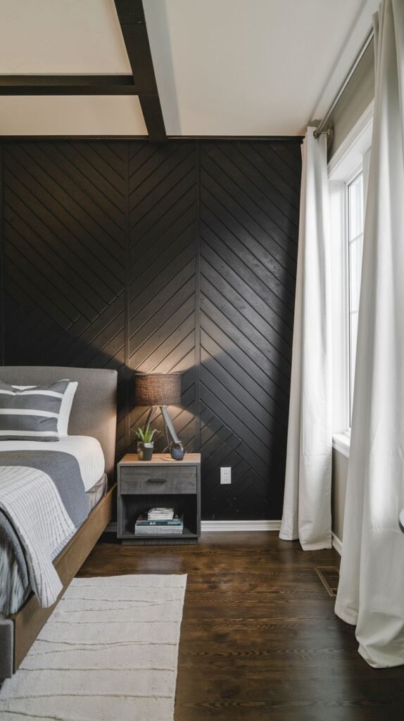 A close-up of a dark, rustic-modern bedroom featuring a full accent wall covered in dark wood planks arranged in a distinct chevron or herringbone pattern.