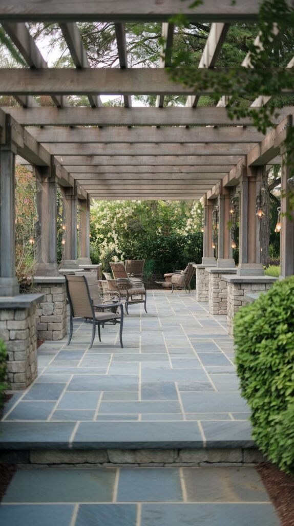 Formal wooden pergola corridor covering a walkway paved with large blue-gray flagstones, supported by columns resting on stacked stone piers, and illuminated by hanging globe lights.