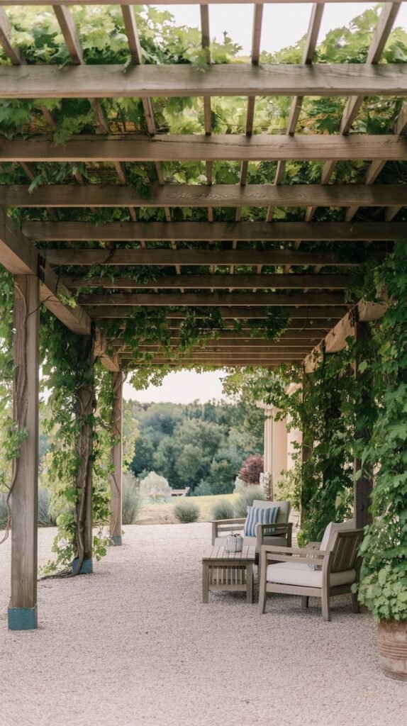 Open pergola with heavy vine coverage shading a seating area with two wooden lounge chairs, set upon a light crushed gravel floor.