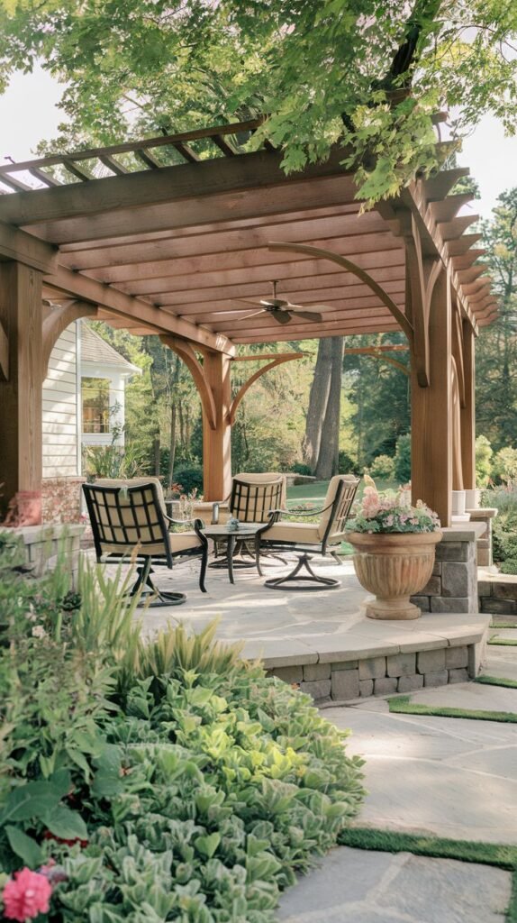 Architecturally detailed wooden pergola featuring curved supports and a ceiling fan, covering an elevated stone patio furnished with four cushioned swivel lounge chairs and accented by a classical stone urn planter.
