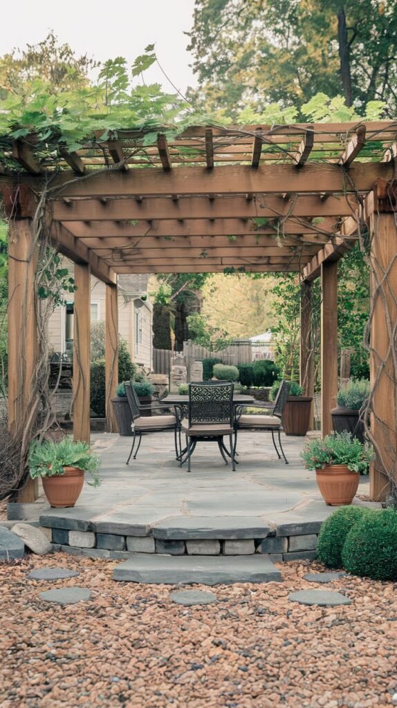 Elevated stone patio with a wooden pergola heavily draped in green vines, furnished with a dark metal dining set, and bordered by stone steps leading down to a gravel path with stepping stones.