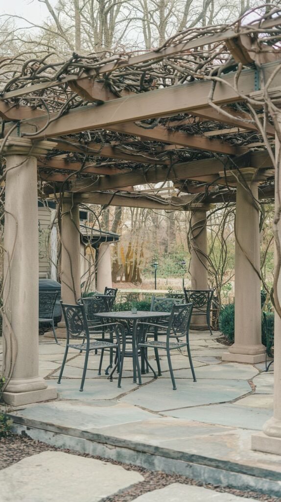 Classical pergola supported by round columns, featuring a canopy woven from bare vines, sheltering a black metal dining set on a patio paved with large, irregular flagstones.