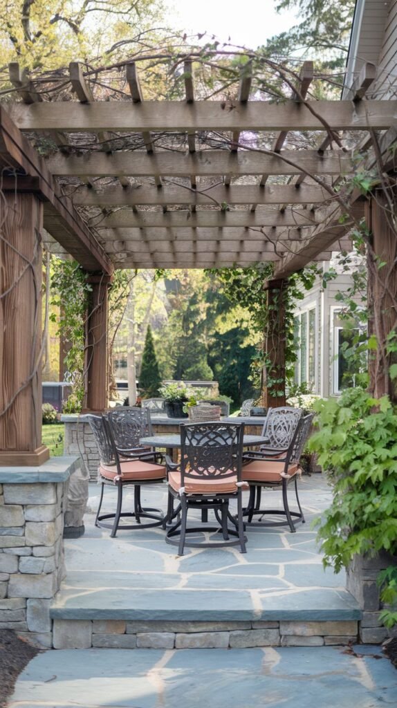Wooden pergola structure with vine accents over an elevated patio made of irregular blue-gray flagstone, supported by stacked stone bases, and furnished with a dark metal dining set.