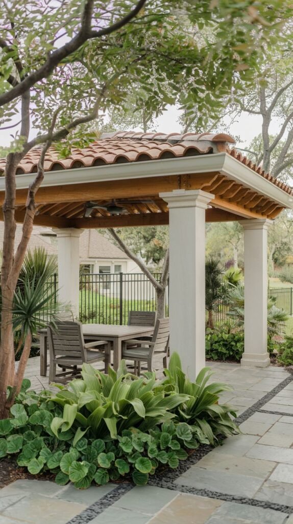 Mediterranean-style covered pavilion featuring a terracotta tile roof and white square columns, sheltering a wooden dining set on a blue-gray flagstone patio outlined with dark pebbles.