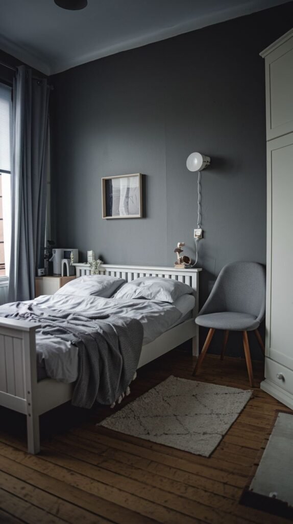 Monochrome bedroom with dark gray walls and curtains, a white slatted bed frame, an adjacent light gray upholstered chair, and a hint of a pale green wardrobe.