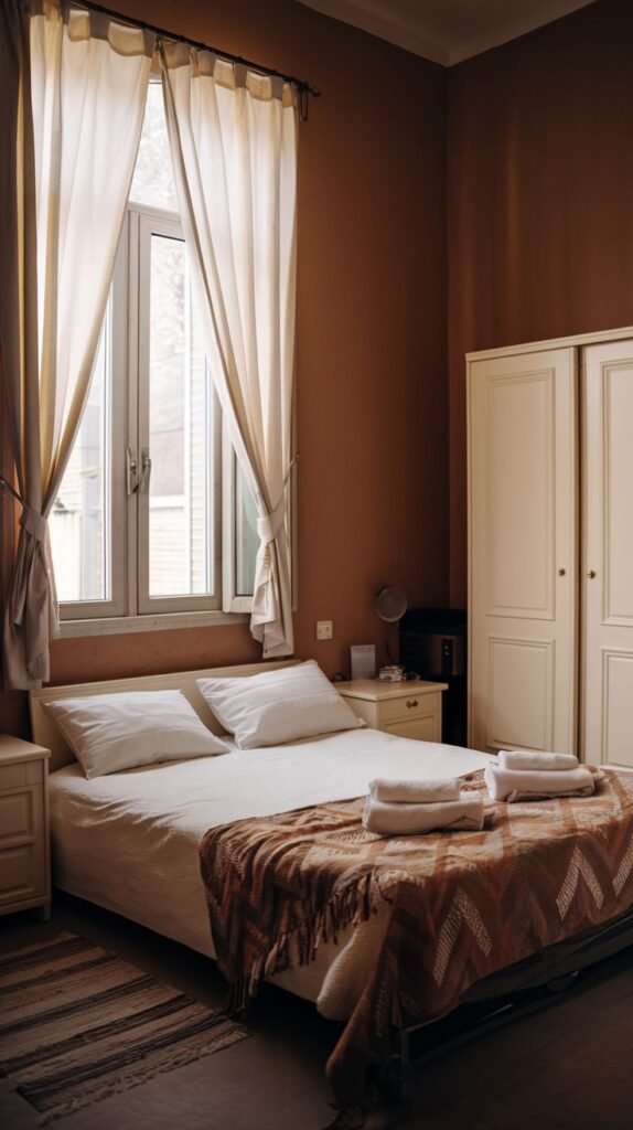 Bedroom with warm reddish-brown walls, a large window draped with voluminous sheer curtains, and a contrasting large white freestanding wardrobe.