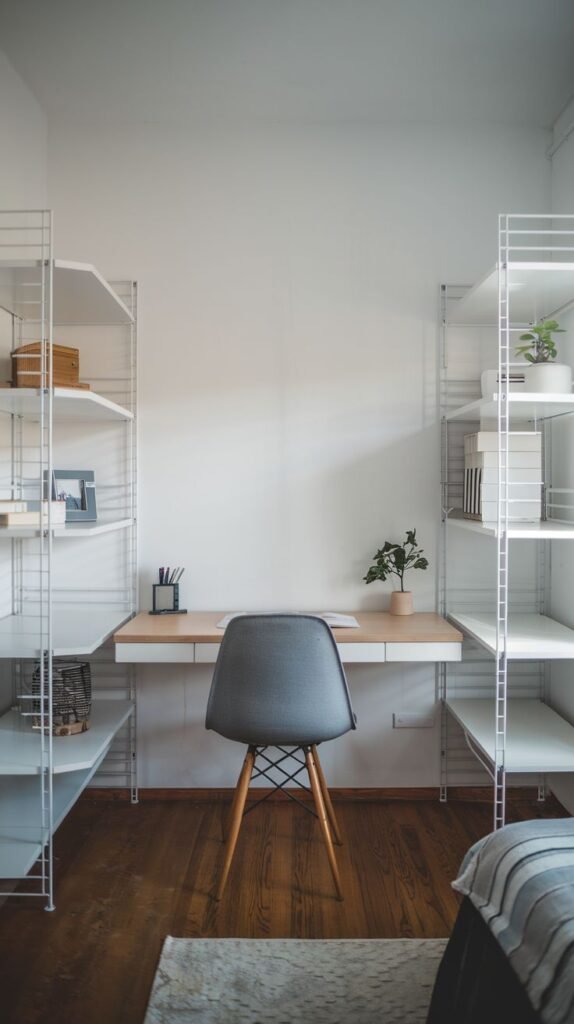 A clean, modern workspace centered around a light wood desk surface suspended between two tall, white wire-frame modular shelving units that line the white walls. A gray fabric chair sits at the center, facing the desk.