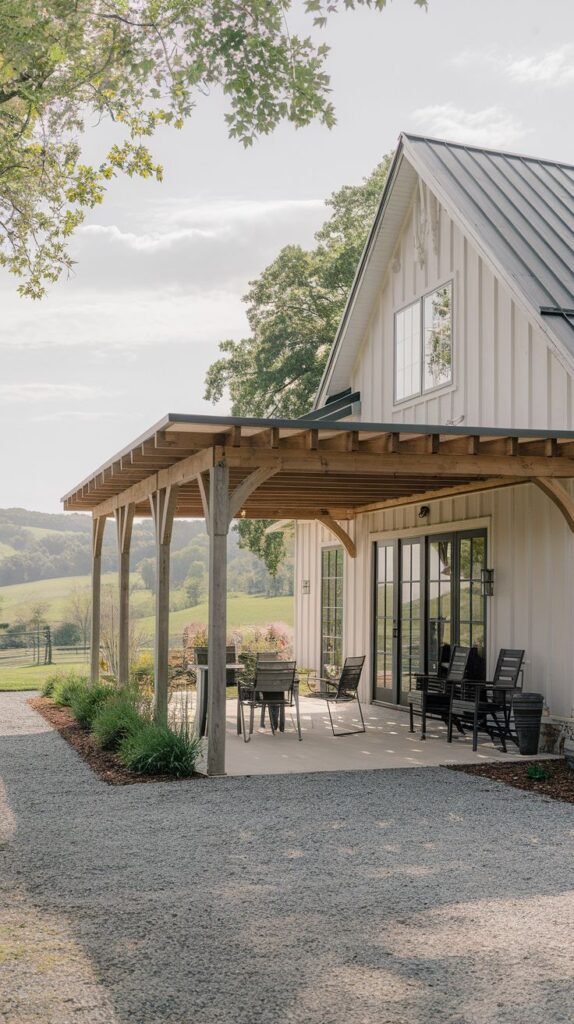 Covered concrete patio attached to a white vertical board house, featuring dark modern furniture, shaded by a shed roof supported by unique rounded, natural wooden posts, bordered by gravel.