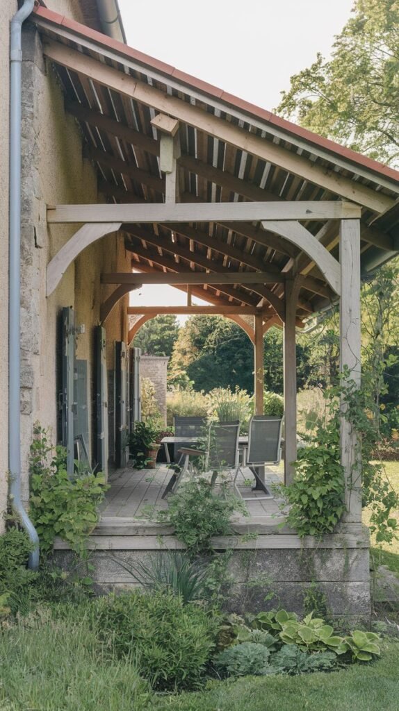 Covered wooden deck porch featuring distinct arched timber beams and posts with exposed rafters, surrounded by lush ground landscaping, attached to a textured, stucco or stone house exterior.