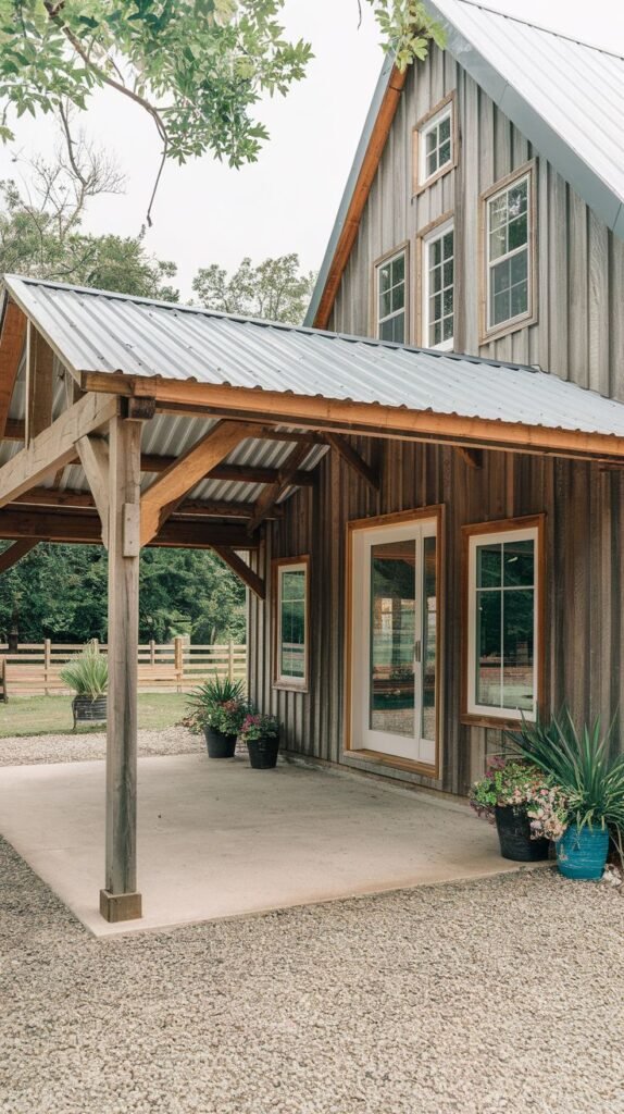 Covered concrete patio with a simple corrugated metal shed roof and basic wood supports, attached to a house clad in gray, weathered vertical wood siding, bordered by gravel.