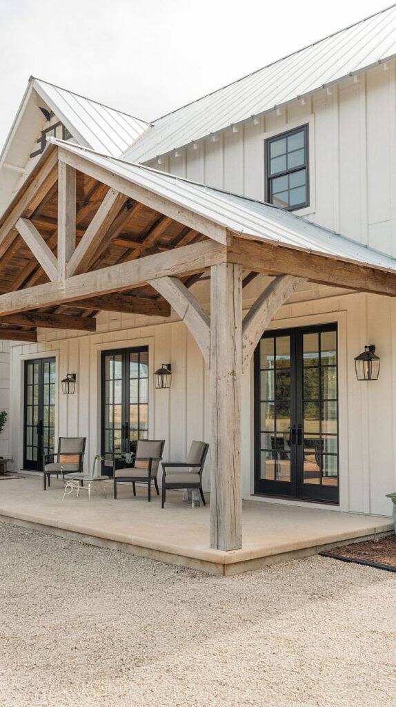 Wide concrete entry patio featuring a massive exposed timber gable roof structure supported by large central posts, attached to a white vertical board house with black-framed French doors, adjacent to a gravel drive.