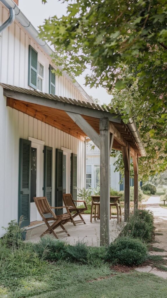 Long covered wooden porch with a corrugated metal shed roof and round wood posts, shading a path, running along a white vertical board house with dark green shutters.