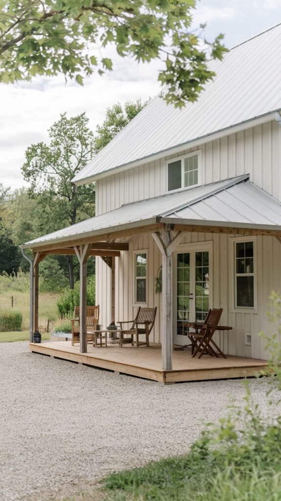 Long covered wooden deck porch with a simple shed roof supported by wooden posts, attached to a white vertical board house, located next to a gravel driveway.