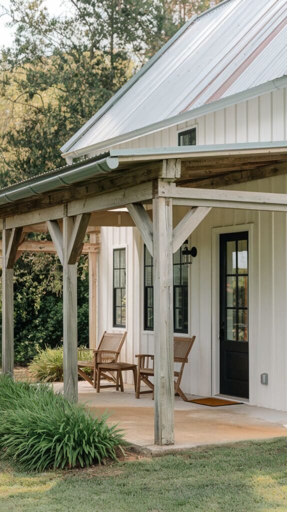 Small covered concrete patio with a sloped corrugated metal roof supported by rustic wood posts, adjacent to a white vertical board house with a black-framed door, featuring woven wicker chairs.