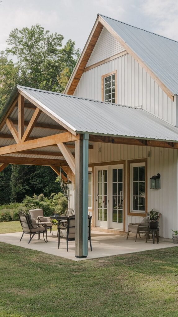 Gabled covered patio extension featuring exposed wood trusses and a corrugated metal roof, shading a concrete slab with outdoor seating, attached to a white vertical board house via white French doors.