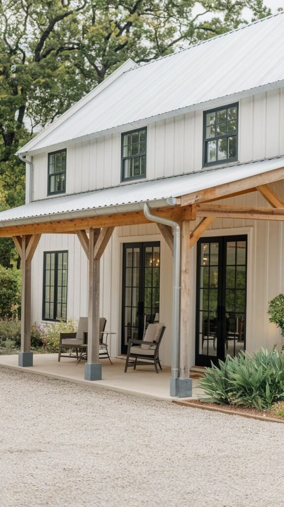 Covered concrete porch patio featuring a sloped roof supported by heavy timber posts, attached to a modern white board-and-batten house with black-framed windows and doors, bordered by a gravel drive.