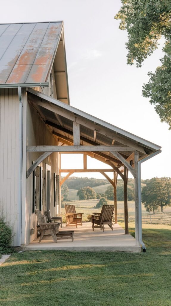 Rustic timber frame covered porch with curved posts and exposed rafters over a concrete floor, featuring wooden Adirondack chairs, attached to a light-sided house with a rusted metal roof, overlooking rolling hills.
