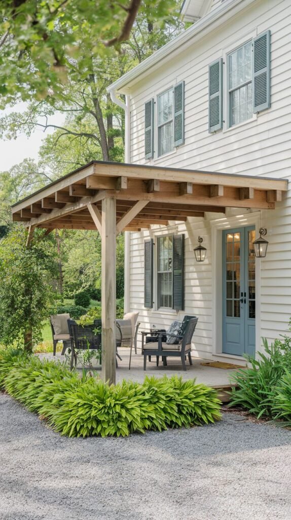 Small covered seating area with a simple sloped wooden lean-to roof structure, over a concrete spot leading to a gravel patio bordered by lush hosta plants, attached to a white clapboard house with dark shutters.