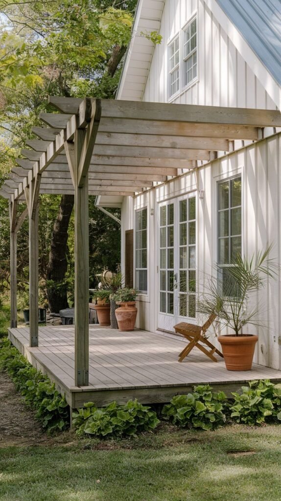 Raised wooden deck patio featuring an open wooden pergola structure providing partial cover, decorated with large terracotta planters, attached to a white vertical board house with white French doors.