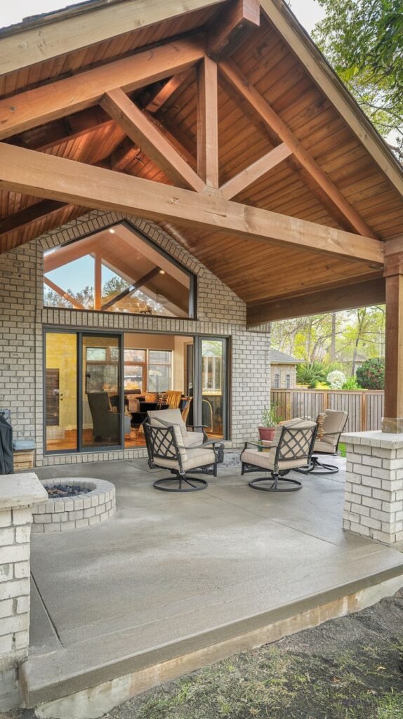 Gabled covered patio with exposed wood trusses and a finished wood ceiling, attached to a light brick house. The concrete patio floor is lined with low brick walls, centering four swivel chairs around a built-in circular fire pit.