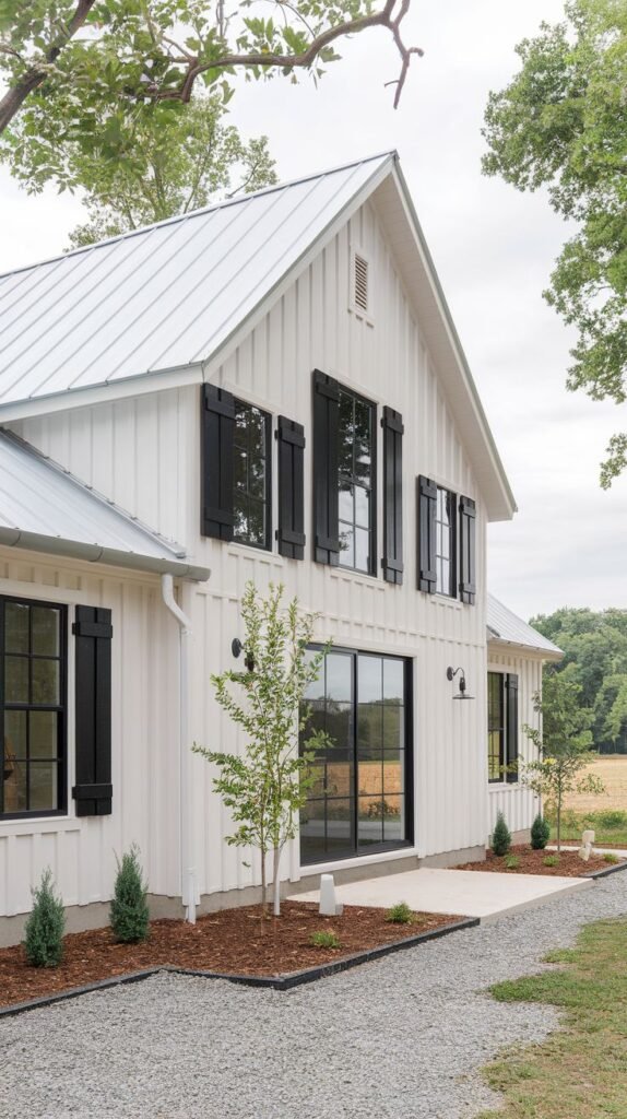 White farmhouse with a silver metal roof. Upper windows have black board-and-batten shutters, while the lower level features a large, black-framed sliding glass door leading to a concrete patio and gravel path.