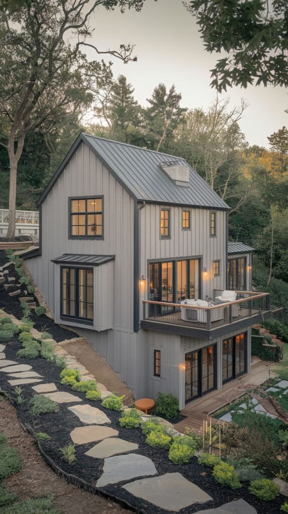 Dramatic multi-level dark grey farmhouse built on a slope, with a grey metal roof. The mid-level features a wooden balcony deck secured by a black railing, and access below is via large, irregular stone steps set into the landscaped hillside.